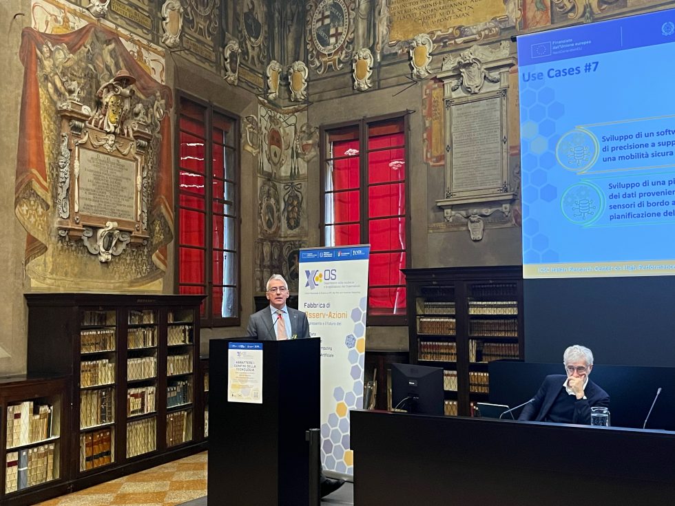 Immagine che raffigura un relatore al podio all'interno di una sala storica affrescata con librerie antiche. Alle sue spalle, un moderno schermo proietta una slide blu intitolata "Use Cases #7" con icone relative a sviluppo software e mobilità. Un'altra persona è seduta ad ascoltare sulla destra.