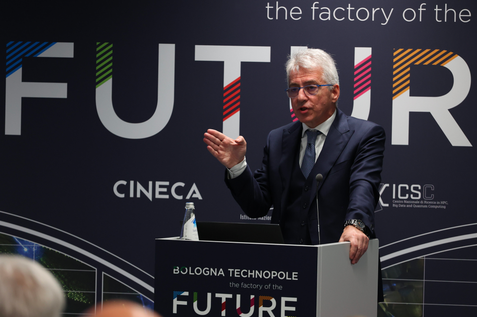 Gemini said Ecco la descrizione ottimizzata come testo alternativo per l'immagine indicata: Immagine che raffigura un relatore mentre tiene un intervento dietro a un podio durante un evento istituzionale. L'uomo, che indossa un abito scuro, occhiali e cravatta, è colto nell'atto di parlare gesticolando con la mano destra. Sul podio è visibile il marchio "Bologna Technopole - the factory of the Future", mentre il fondale scuro alle sue spalle riporta a grandi caratteri la scritta "FUTURE" insieme ai loghi del CINECA e del centro ICSC