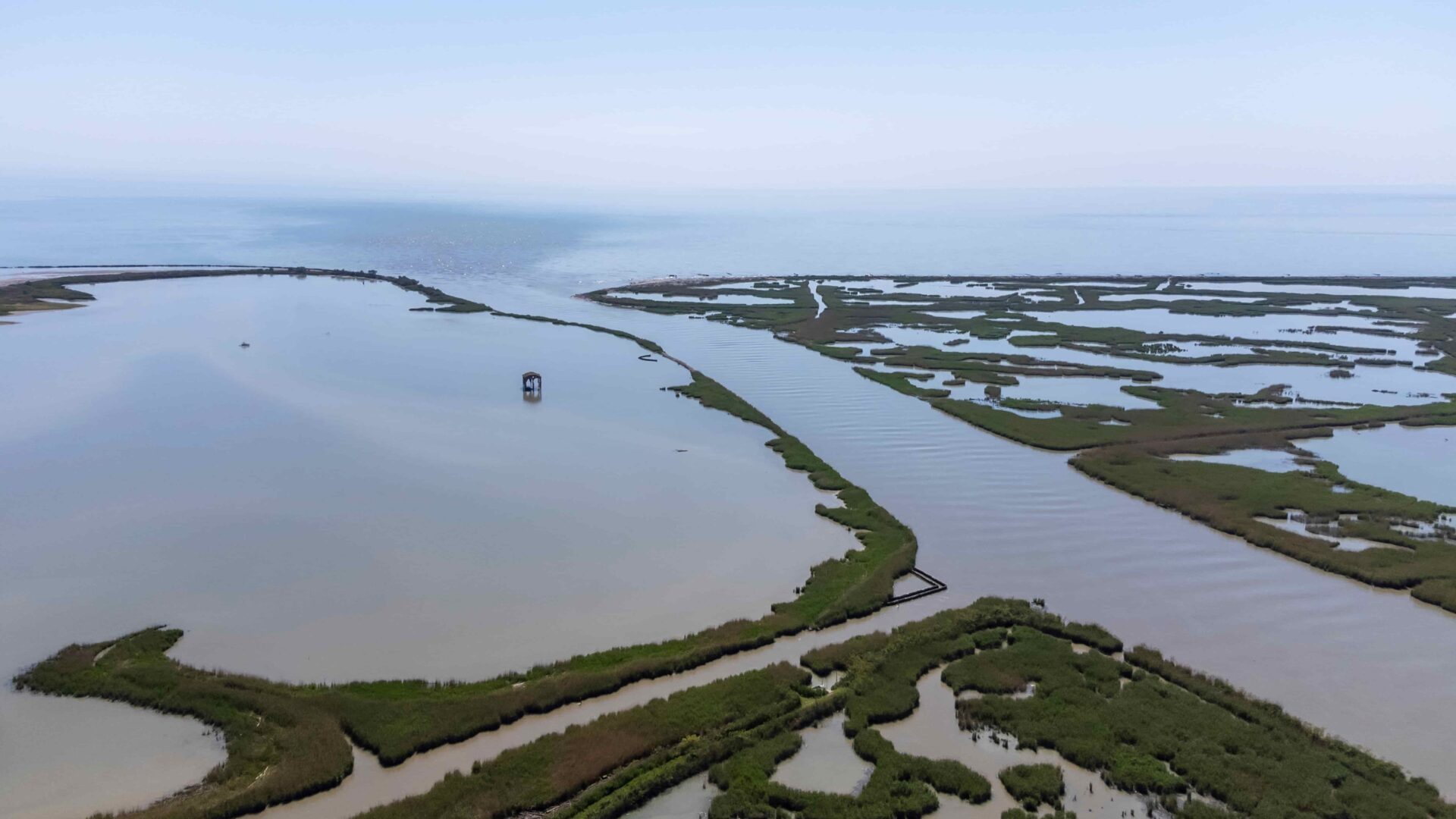 immagine che rappresenta una veduta aerea di un paesaggio lagunare o del delta di un fiume, in cui l'acqua dolce incontra il mare aperto sullo sfondo. L'area è caratterizzata da ampi specchi d'acqua calma e da un intricato sistema di lingue di terra e isolotti ricoperti di fitta vegetazione verde e canneti. Sulla sinistra, nel bacino d'acqua più grande, spicca una piccola struttura isolata su palafitte, mentre sulla destra la foce è solcata da un canale principale circondato da zone paludose sotto un cielo chiaro e leggermente fosco.